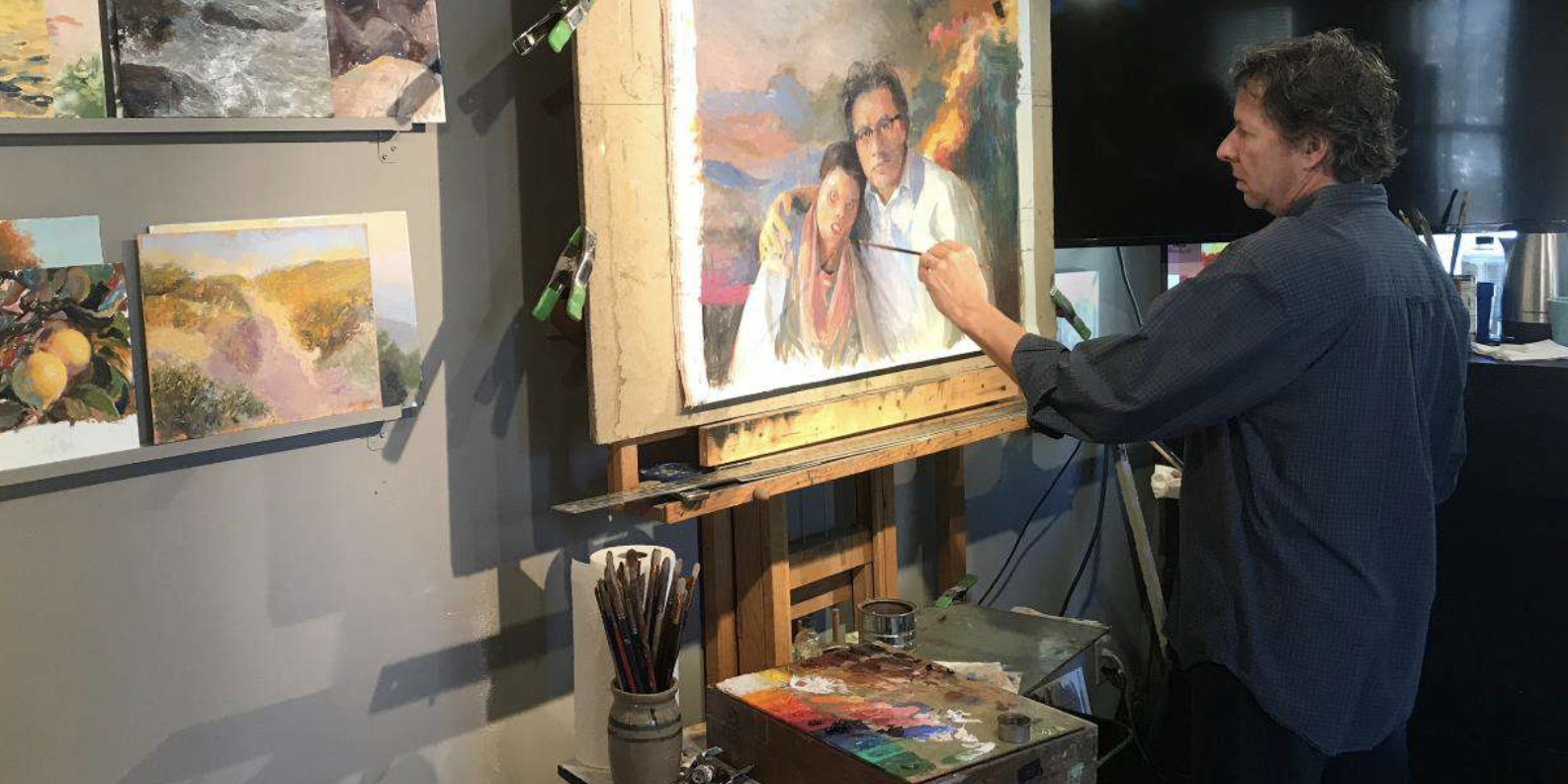 Artist Timothy Chambers stands at his easel painting a portrait of a man with his patient, a young woman who is a burn victim. In the foreground are a palette of oil paints and oil painting brushes.
